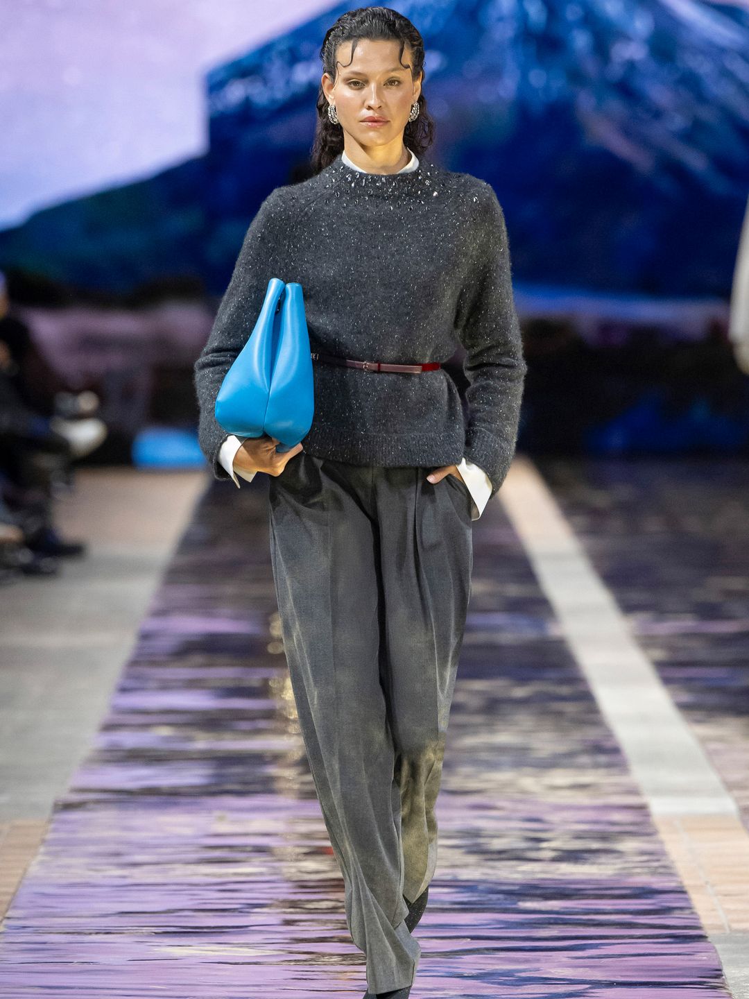 A model walks the runway at the Marc Cain show wearing a grey jumped, belted with a slim burgundy belt, loose fit grey trousers and she is carrying a large bright blue clutch bag. 