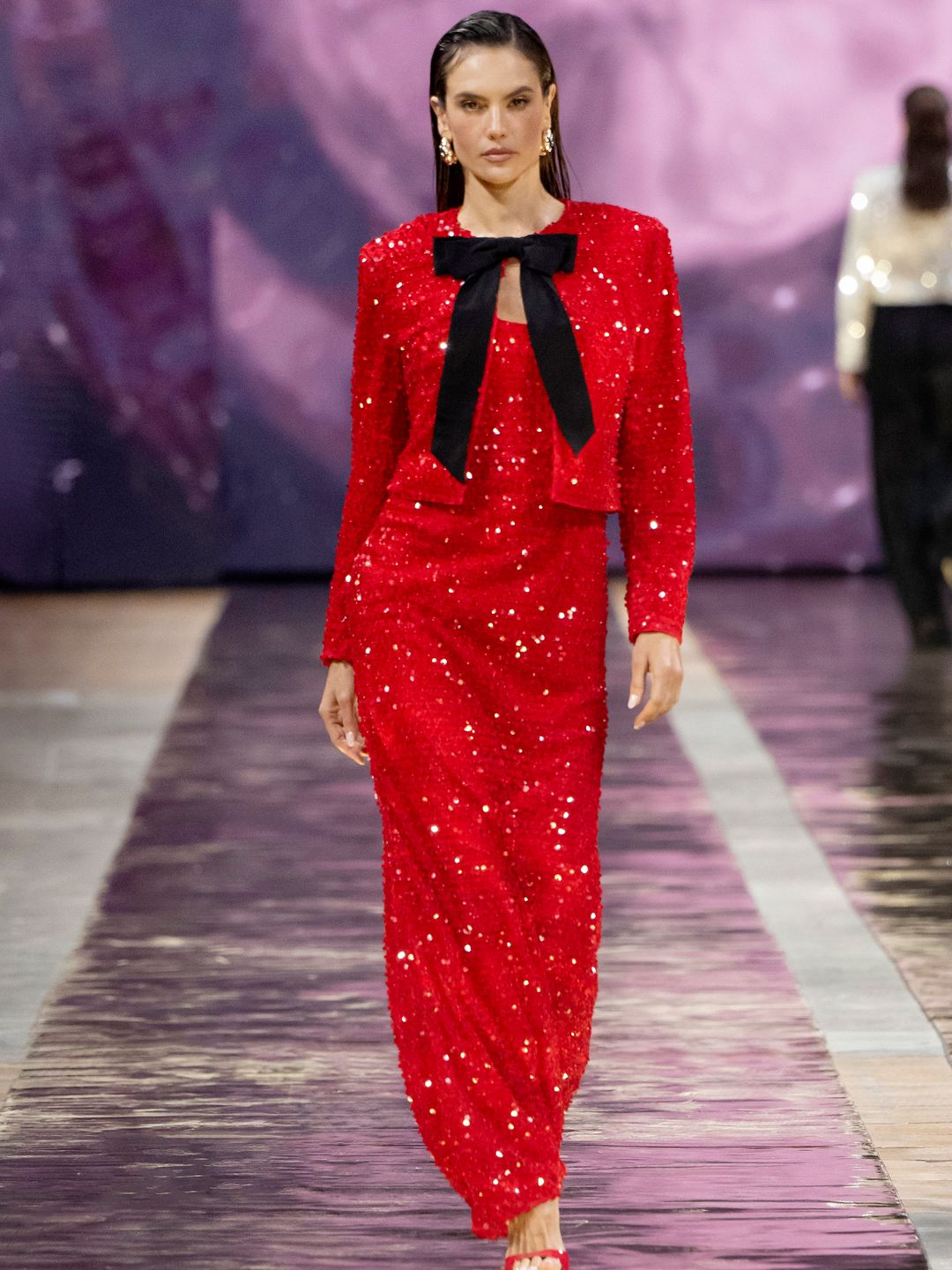 Alessandra Ambrosio walks the Marc Cain runway in a floor length bright red sequin dress with matching jacket with large black bow detail on the front. She is also wearing red peeptoe heels, long gold earrings and her hair is slicked back