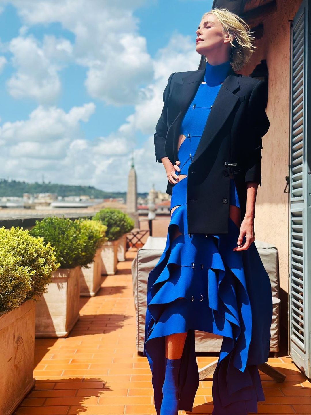 Charlize posing in her blue dress with a black blazer behind her is a vista of Rome