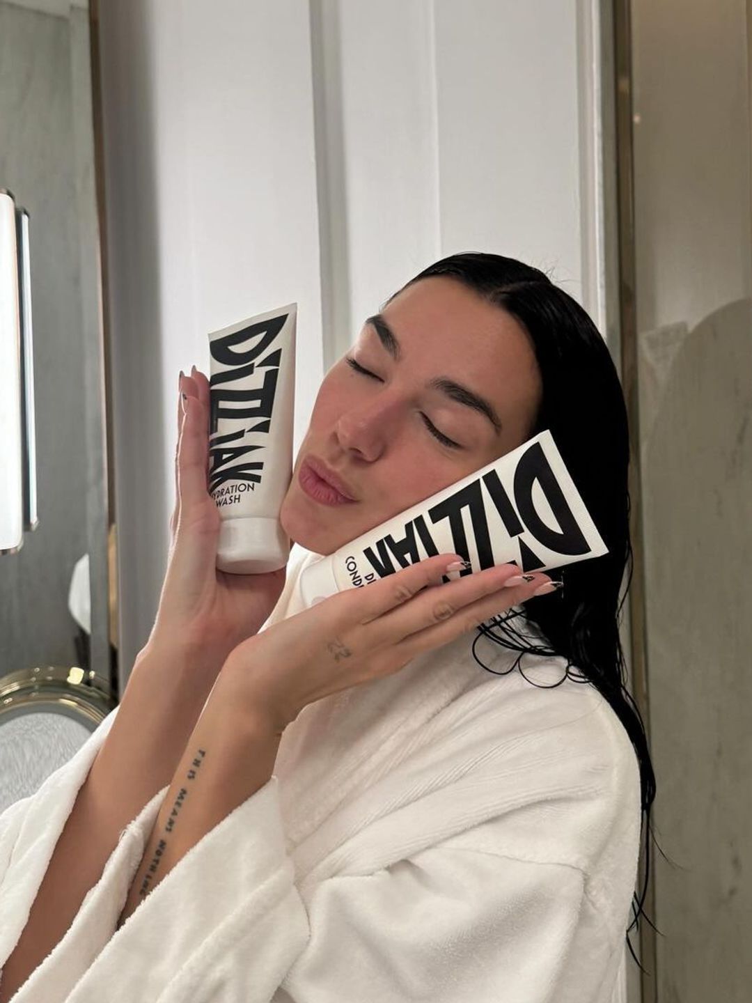 Dua Lipa, wearing a white bathrobe with wet hair, gently presses two Dizziak haircare products against her cheeks with a serene, closed-eyed expression in a bathroom setting.