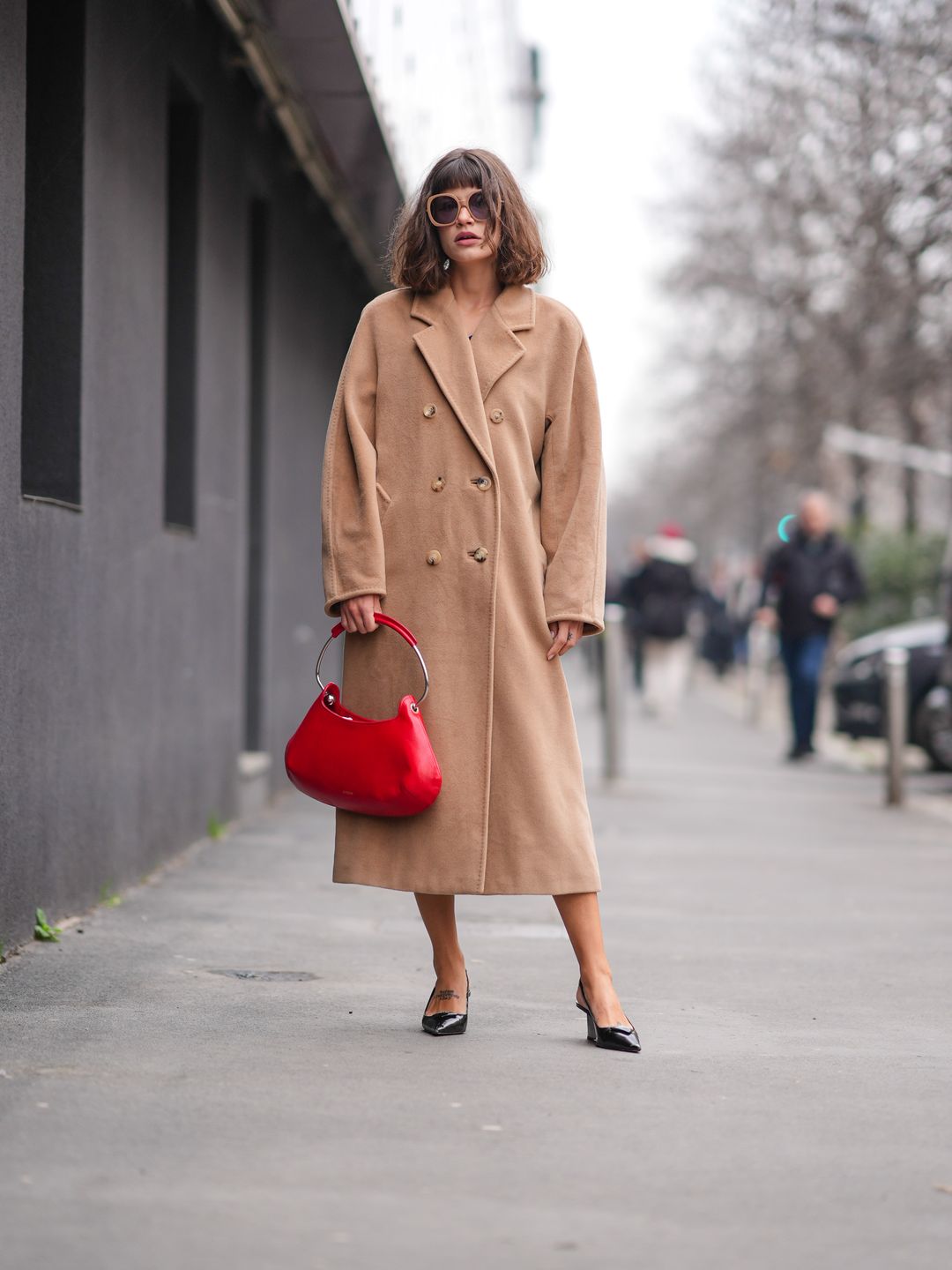 A guest wears sunglasses, a beige double breasted long winter coat, a red leather bag, black pointed shoes , outside Max Mara