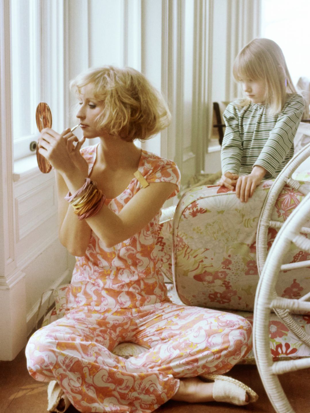 Model Chris Royer seated on the floor in front of window applying makeup with a handheld mirror, a child standing behind her and holding up a seat cushion. She is wearing Lilly Pulitzer's orange and white bird-patterned Dacron polyester and cotton pajama set of capsleeve T-shirt and pants. Accessories: bangles by Lecour-Johnson. Makeup by Sandra Linter. Hair by Harry King of Cinandre.