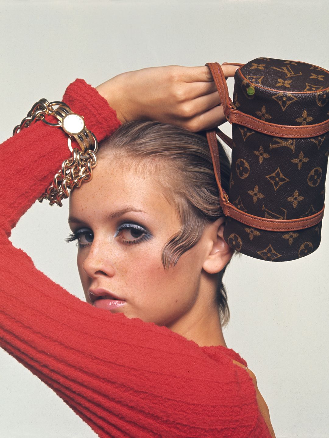 Twiggy poses in a striking red top and showcases a Louis Vuitton monogrammed bag, perfectly styled on her head. Her bold eye makeup and slicked-back hair represent the high-fashion editorial aesthetic, blending luxury accessories with avant-garde styling.