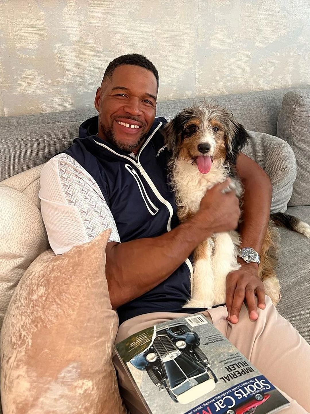 Michael Strahan sat smiling on a couch cradling his pet dog Zuma, also smiling, at his side