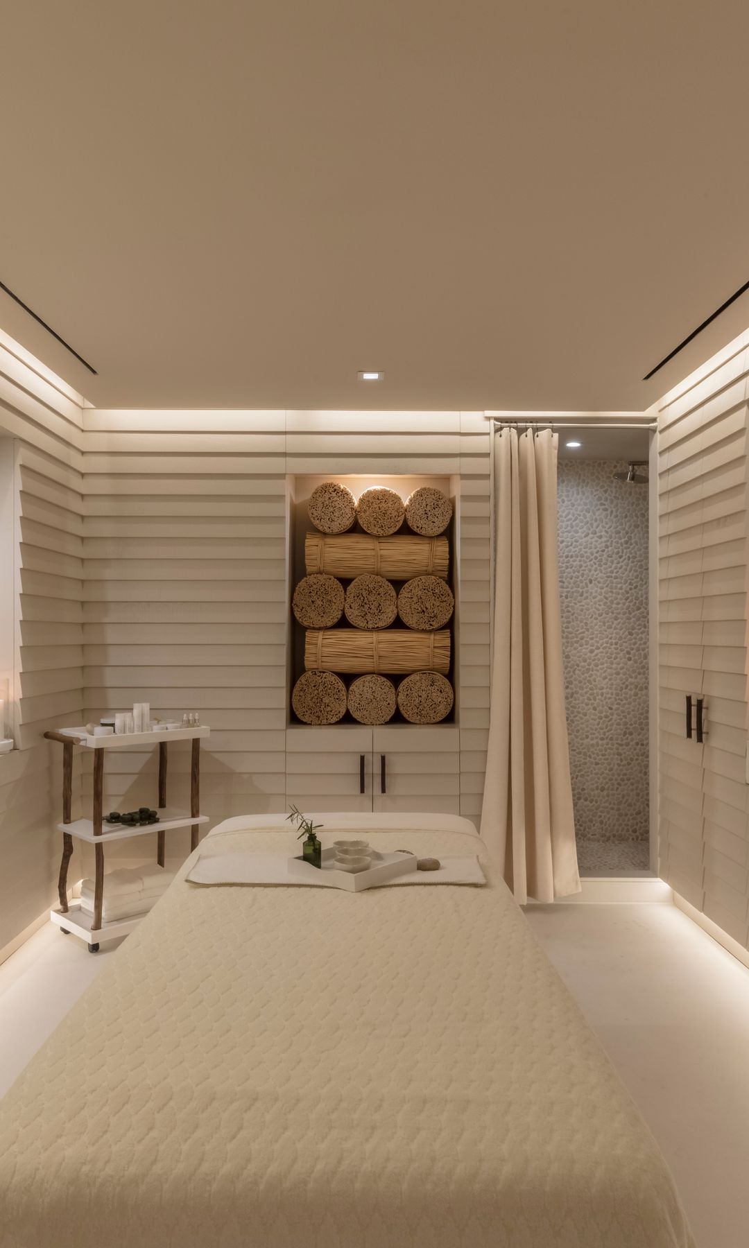 A tranquil, minimalist spa treatment room with warm ambient lighting. A massage bed sits at the center, covered in a beige quilted blanket with a small towel and sprig of greenery placed on top.