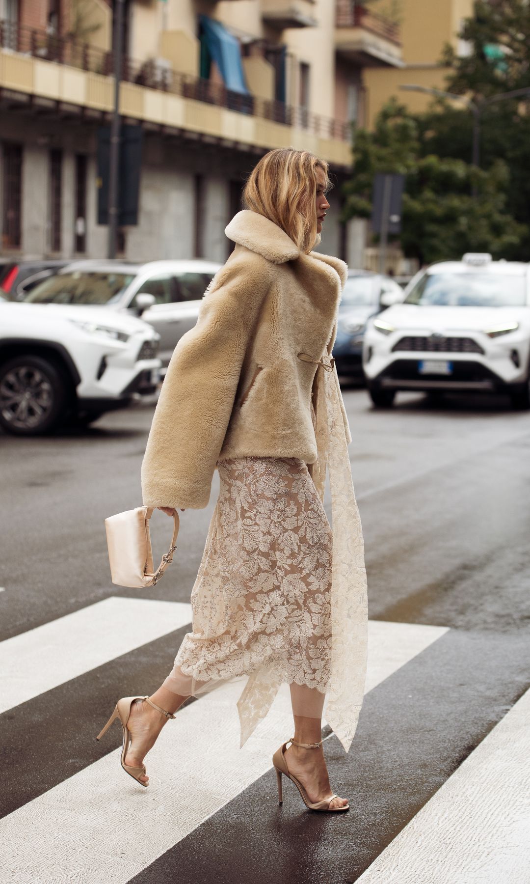 Chloe Lecareux wears a beige shearling coat over a sheer floral lace dress, styled with metallic sandals and a pale pink handbag outside Ermanno Scervino show during the Milan Fashion Week Womenswear Spring/Summer 2026 on September 27, 2025 in Milan, Italy.
