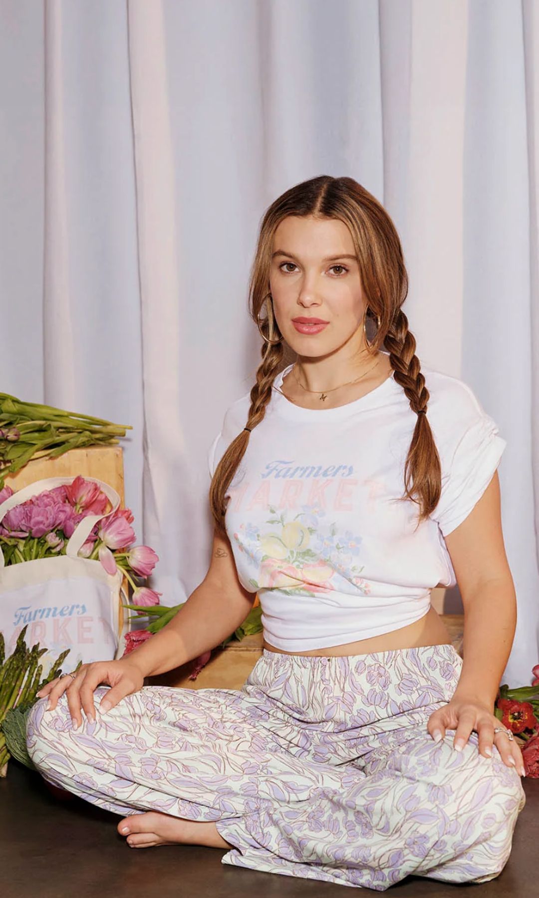 Millie Bobby Brown poses on a crate alongside fresh fruit and vegetables wearing a t-shirt and floral pants