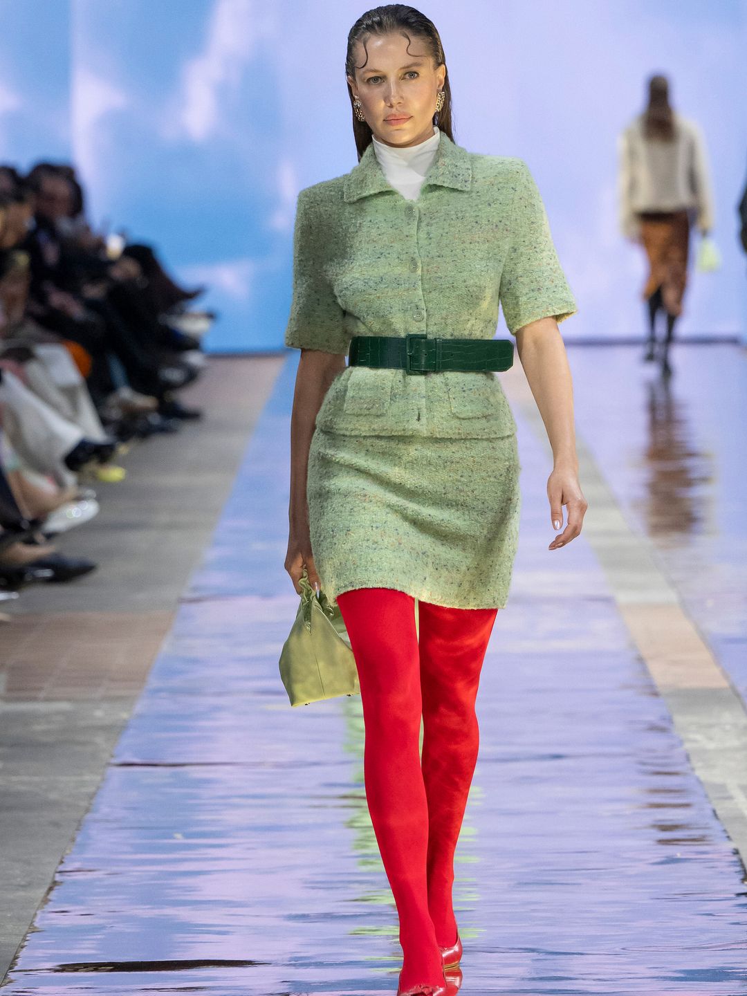 A model walks down the Marc Cain runway in a textured sage green fuzzy mini dress, with a dark green belt. She is also wearing bright red tights and matching shoes and is holding a pale green bag. 