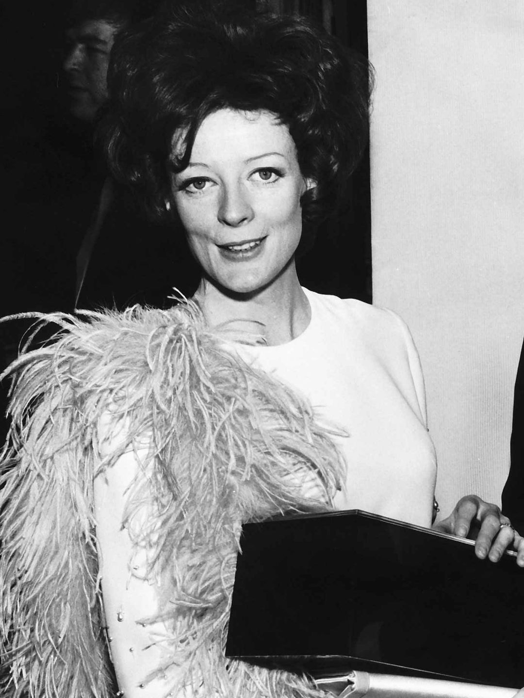 In this image, Maggie Smith is dressed in a glamorous, white gown adorned with feathers. The feathers cover her shoulders and cascade down her arms, adding dramatic flair to her look. She is holding a box, with a soft smile on her face, appearing graceful and poised. The scene captures her in an award ceremony setting, embodying Hollywood glamour with a touch of fun.