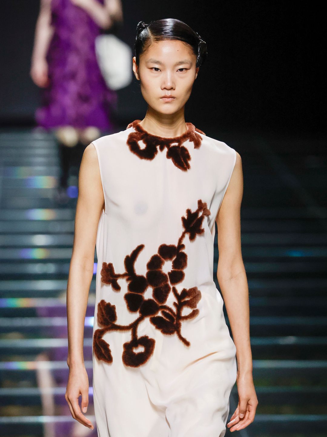A model walks the runway during the Prada Ready to Wear show wearing a white dress with brown floral accents.