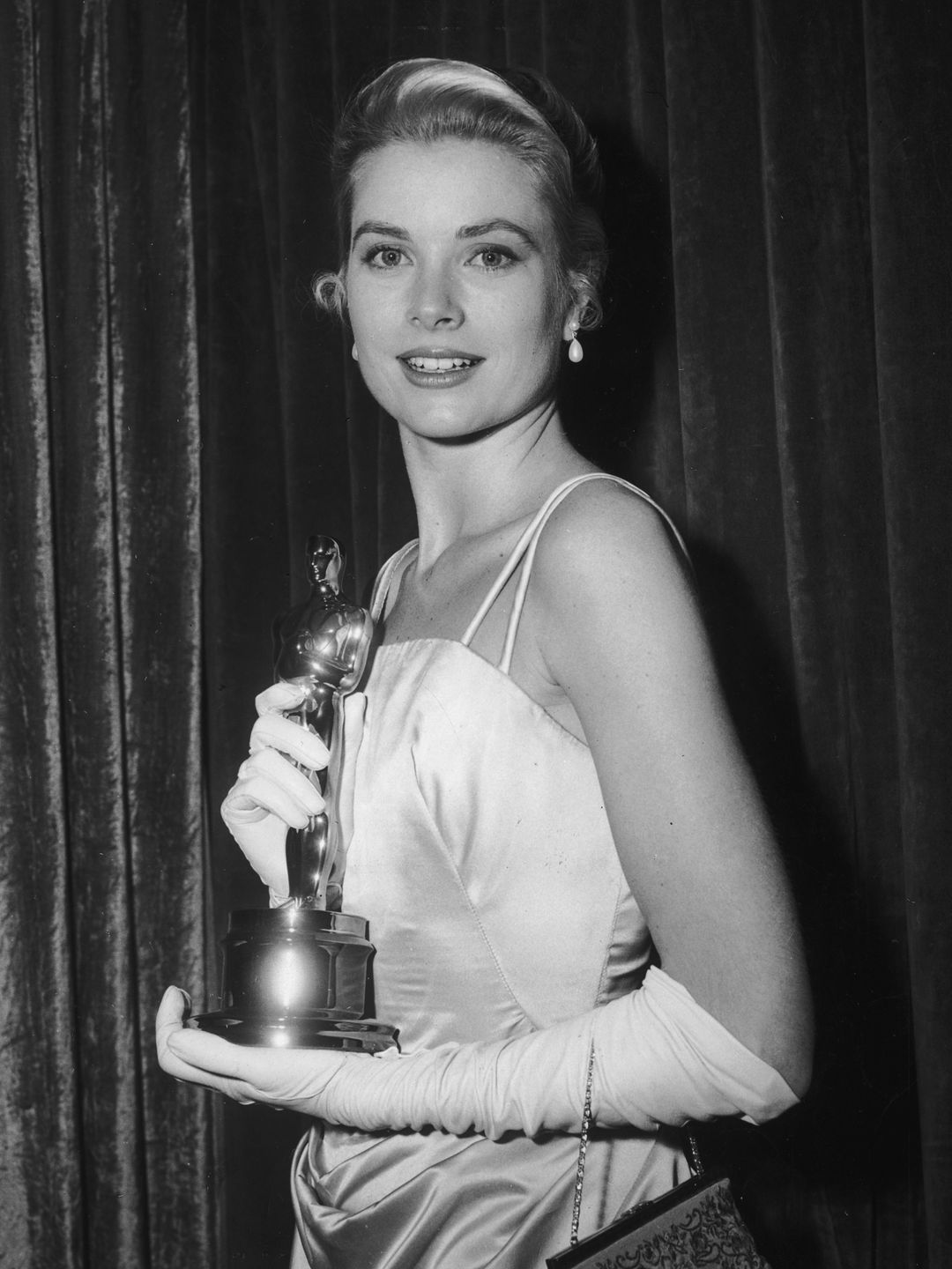30th March 1955:  American actor Grace Kelly (1929 - 1982) poses in front of a curtain with her Best Actress Oscar for director George Seaton's film, 'The Country Girl,' at the Academy Awards, Los Angeles, California. She wears an evening gown with long white gloves.  (Photo by Hulton Archive/Getty Images)