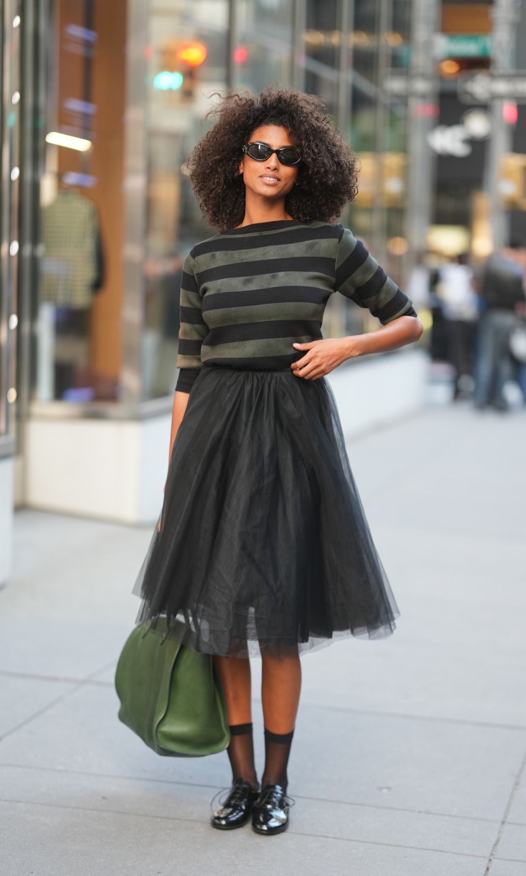 Imaan Hammam is seen wearing a black and olive green striped knit sweater, a Prada black tulle midi skirt, sheer black socks, black oxford shoes, a green tote bag and black sunglasses outside of Ralph Lauren S/S 26 during NYFW