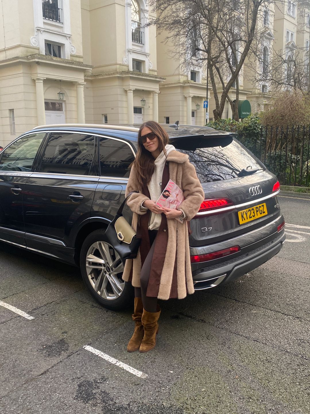Natalie Salmon poses in front of an Audi Q7 in a brown coat and boots for LFW