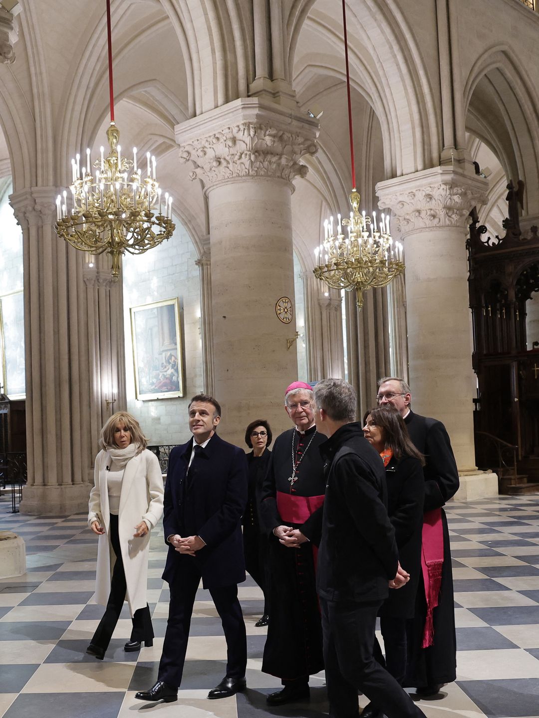 Inside Notre Dame cathedral, Brigitte Macron in a white coat and scarf walks with Emmanuel Macron and others under ornate chandeliers, surrounded by towering columns and checkered floors. They are joined by French Minister of Culture and Heritage Rachida Dati and Paris' archbishop Laurent Ulrich.