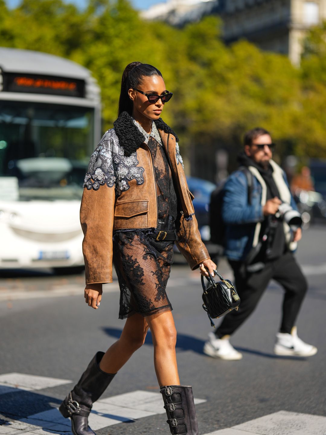 Cindy Bruna wears black sunglasses, diamonds long earrings, a black lace print pattern with diamonds embroidered collar shirt short dress , a brown shiny leather with embroidered crystal shoulder jewels jacket from Miu Miu, a black shiny leather belt, a black embossed stripped pattern handbag from Miu Miu, a lack shiny leather belted knees boots / high boot, outside Miu Miu, during Paris Fashion Week - Womenswear Spring/Summer 2023, on October 04, 2022 in Paris, France. (Photo by Edward Berthelot/Getty Images)