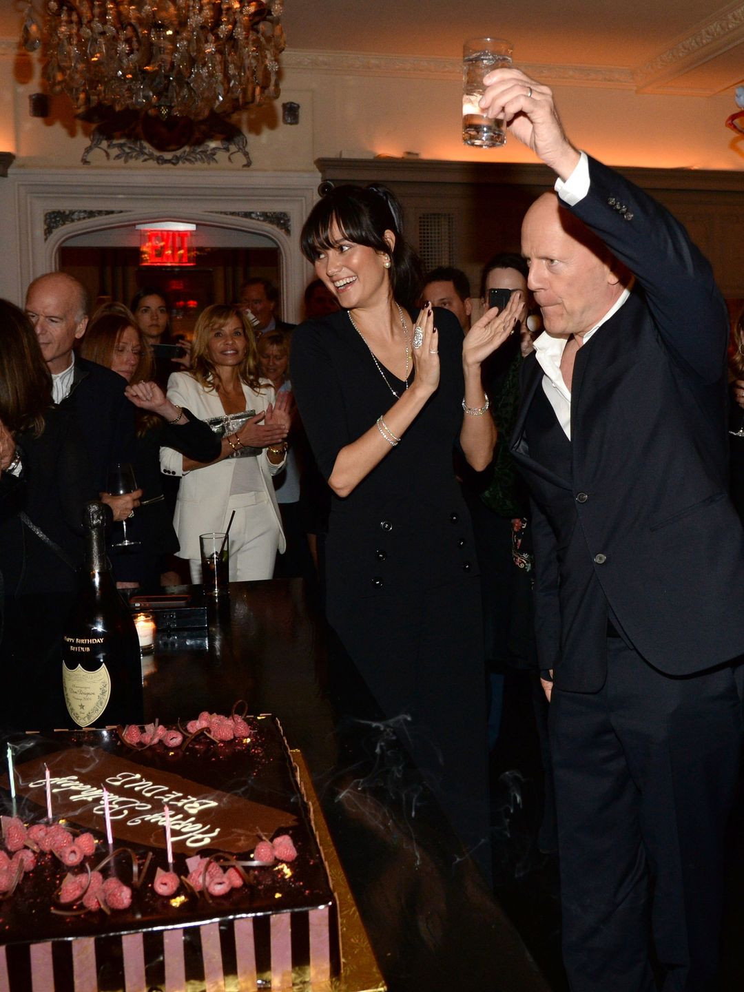 Bruce standing on the right holding up a glass, Emma clapping to his left and to both their left a birthday cake