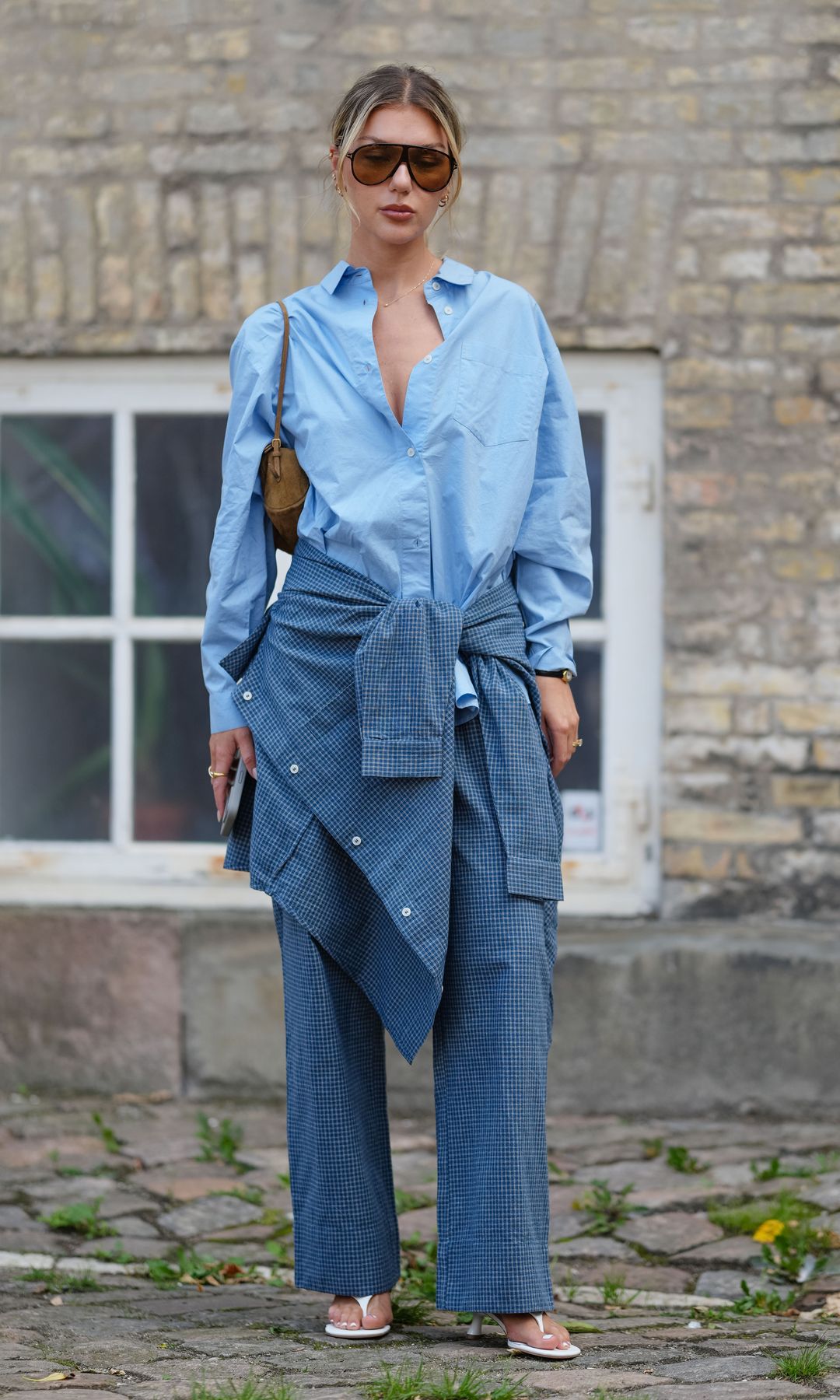  A guest wears a light blue button-up shirt with a relaxed fit and a pointed collar. The sleeves are rolled up slightly. Over the shoulders, a textured blue jacket is tied, featuring visible buttons. The pants are wide-legged and match the jacket in texture and color. Sunglasses with dark lenses and a brown frame are worn. A small brown shoulder bag is carried. Footwear consists of white thong heels sandals. Hair is styled in a low ponytail with loose strands framing the face, outside Skall Studio, during Copenhagen Fashion Week