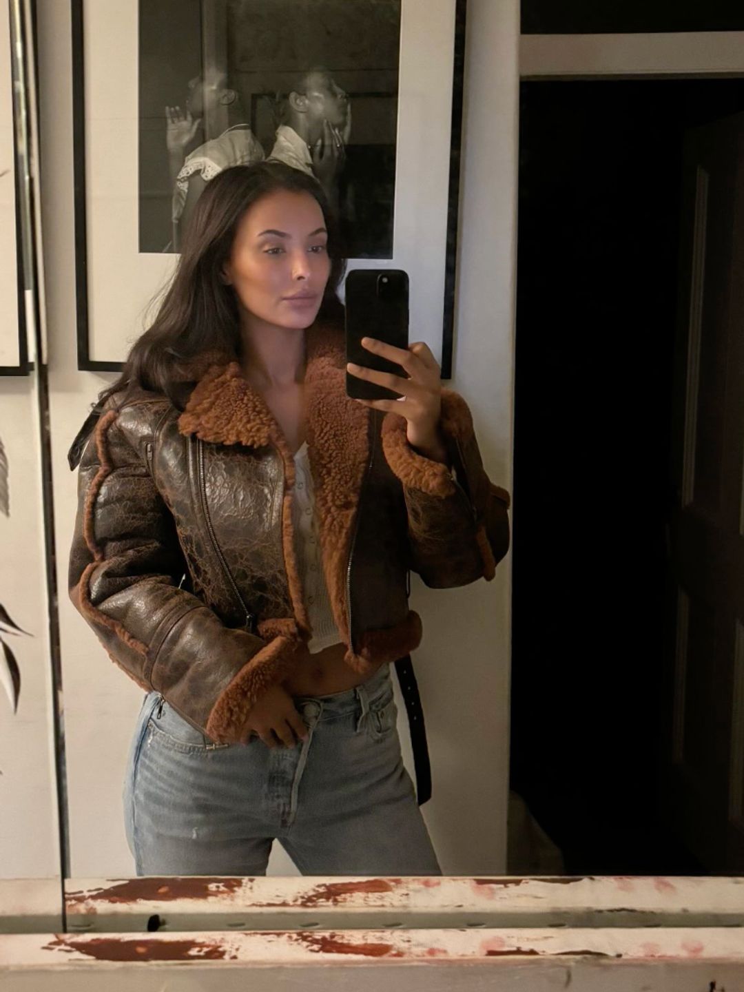 Maya Jama poses for a mirror selfie in brown leather jacket, blue jeans and a white tank top