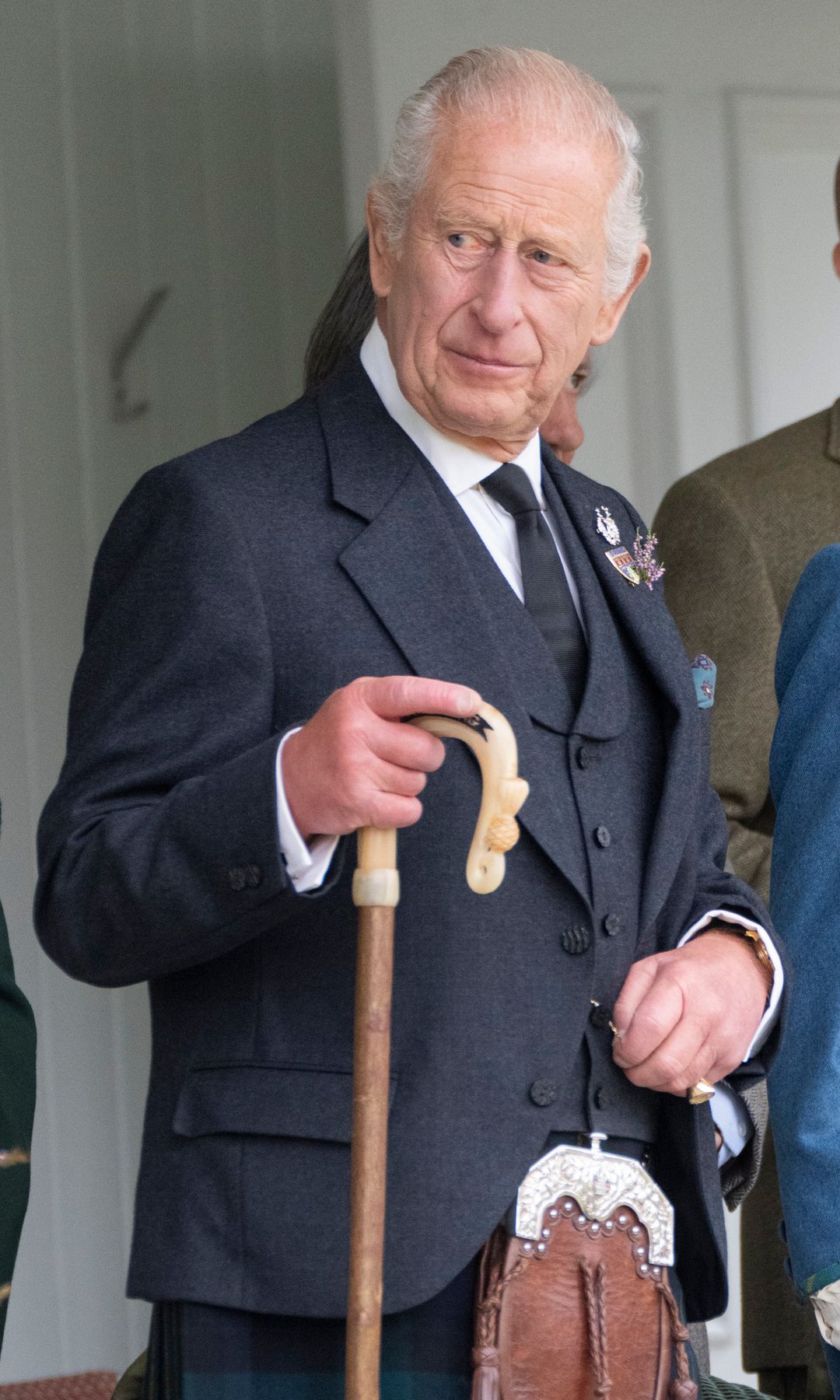 King Charles in a black suit coat and tartan kilt holding a walking stick