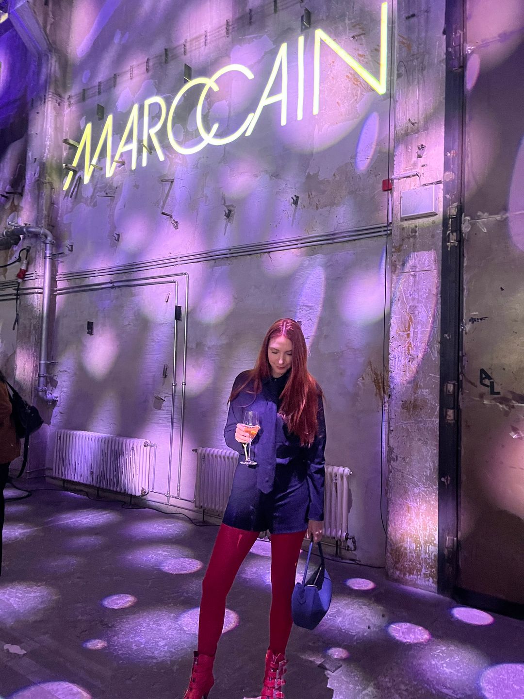 Clare Pennington stands in front of a Marc Cain sign at the Kraftwerk in Berlin. She is wearing a blue pinstripe playsuit with matching tight, red tights and red kitten heel ankle boots with silver buckle detail. She is holding a glass of champagne and a blue handbag,