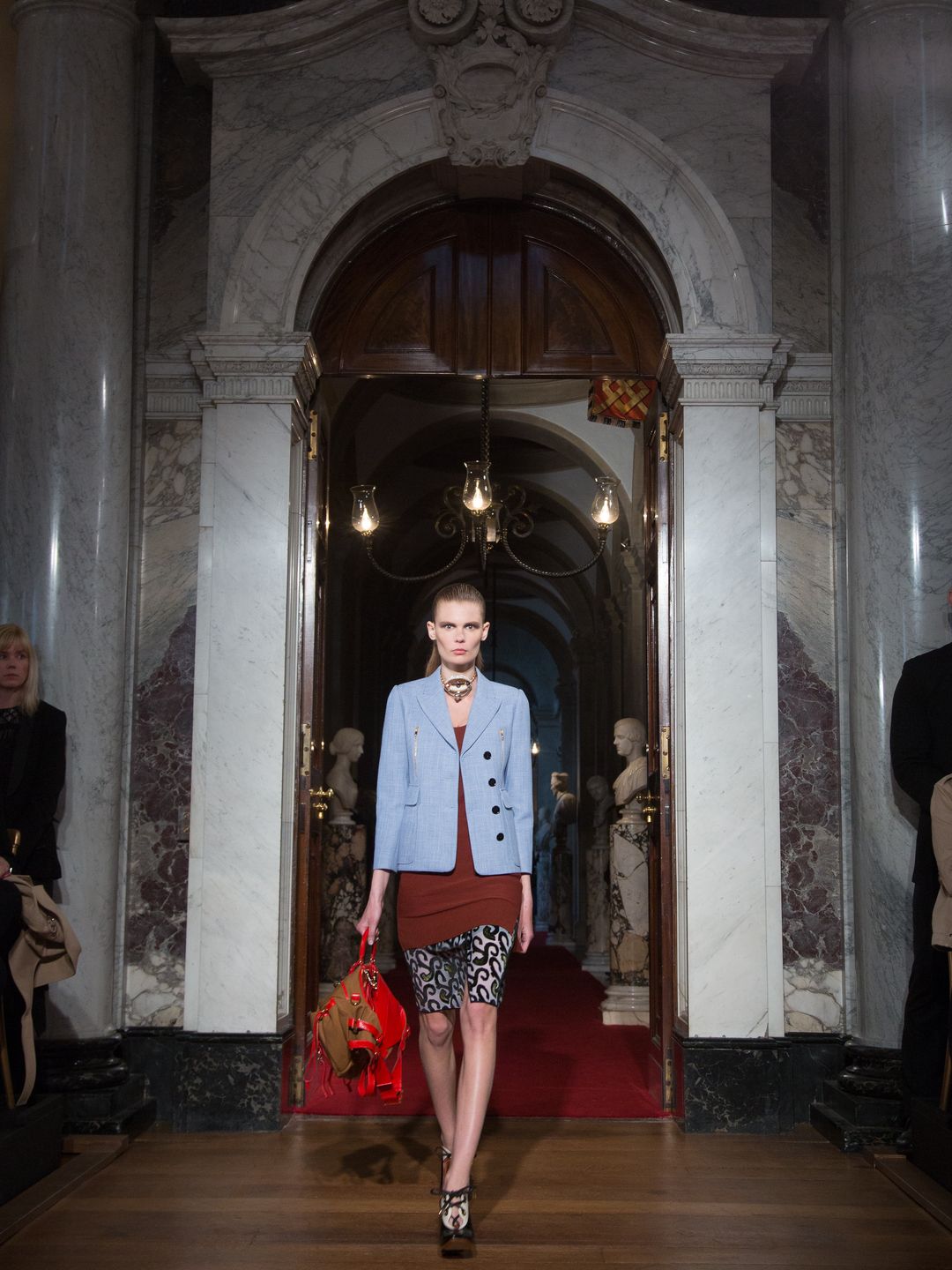 A model walks confidently through a grand marble archway, set in Blenheim Palace’s opulent interiors. Dressed in a chic mix of structured outerwear and patterned leggings, she carries a bold red bag, adding a pop of color to the elegant, muted tones of the palace's marble surroundings.