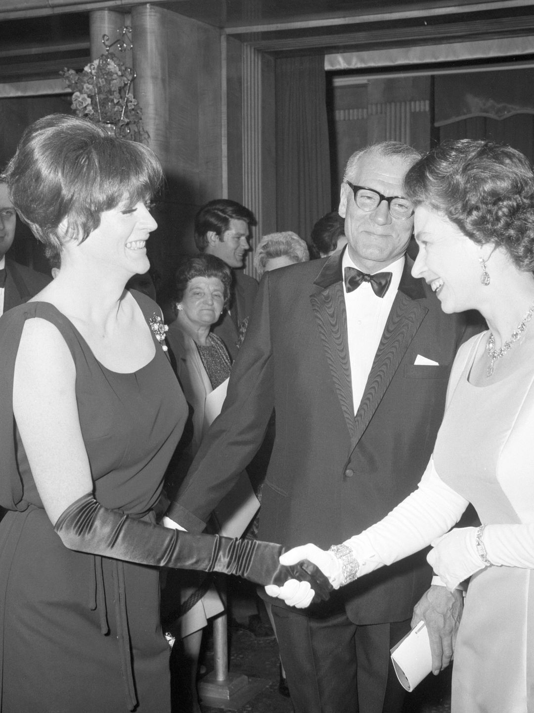 In this black-and-white photo, Maggie Smith is shaking hands with Queen Elizabeth II at a formal event. Maggie wears a sleek black gown and long gloves, her expression warm and smiling as she leans in to greet the Queen. The scene feels regal and momentous, capturing a gracious and poised interaction between a legendary actress and British royalty.