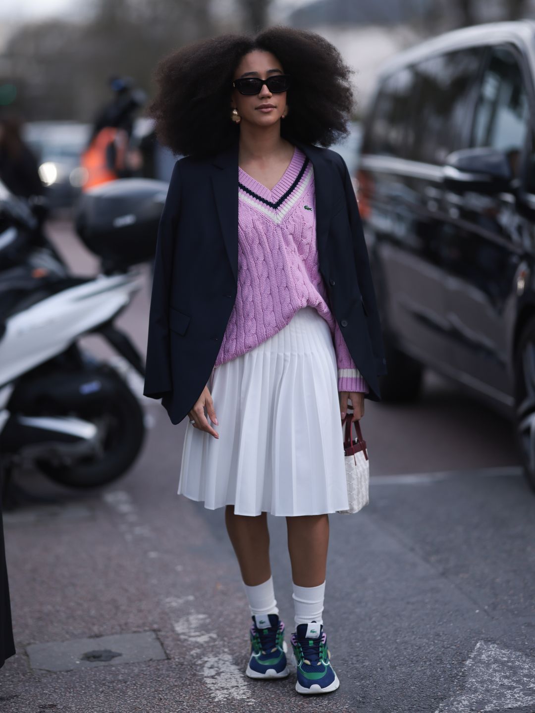 A Guest seen wearing a black blazer jacket, purple Lacoste pullover, white skirt, socks, green sneaker, black shades and white bag outside Lacoste Show during the Womenswear Fall/Winter 2024/2025