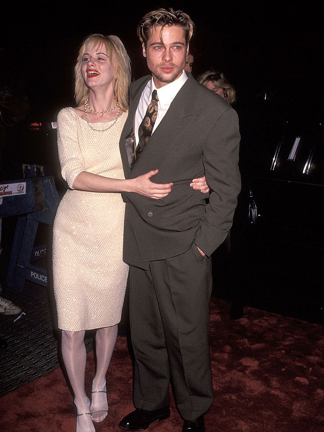 Juliette Lewis cuddling up to Brad Pitt at the Thelma & Louise premiere. She is wearing a gold boat neck dress with shimmering sequins and he is dressed in a grey suit. 