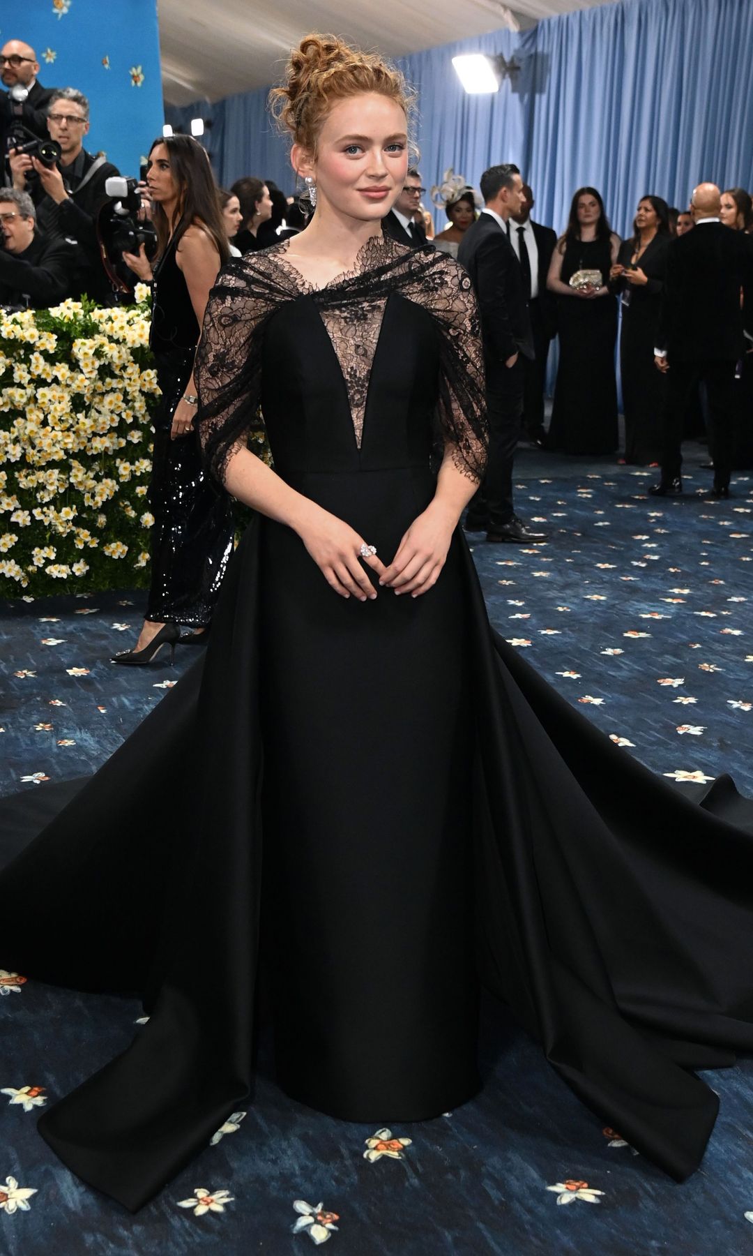 Sadie Sink wears a black dress with train, paired with a short lace black cape and her hair is styled in a bun.
