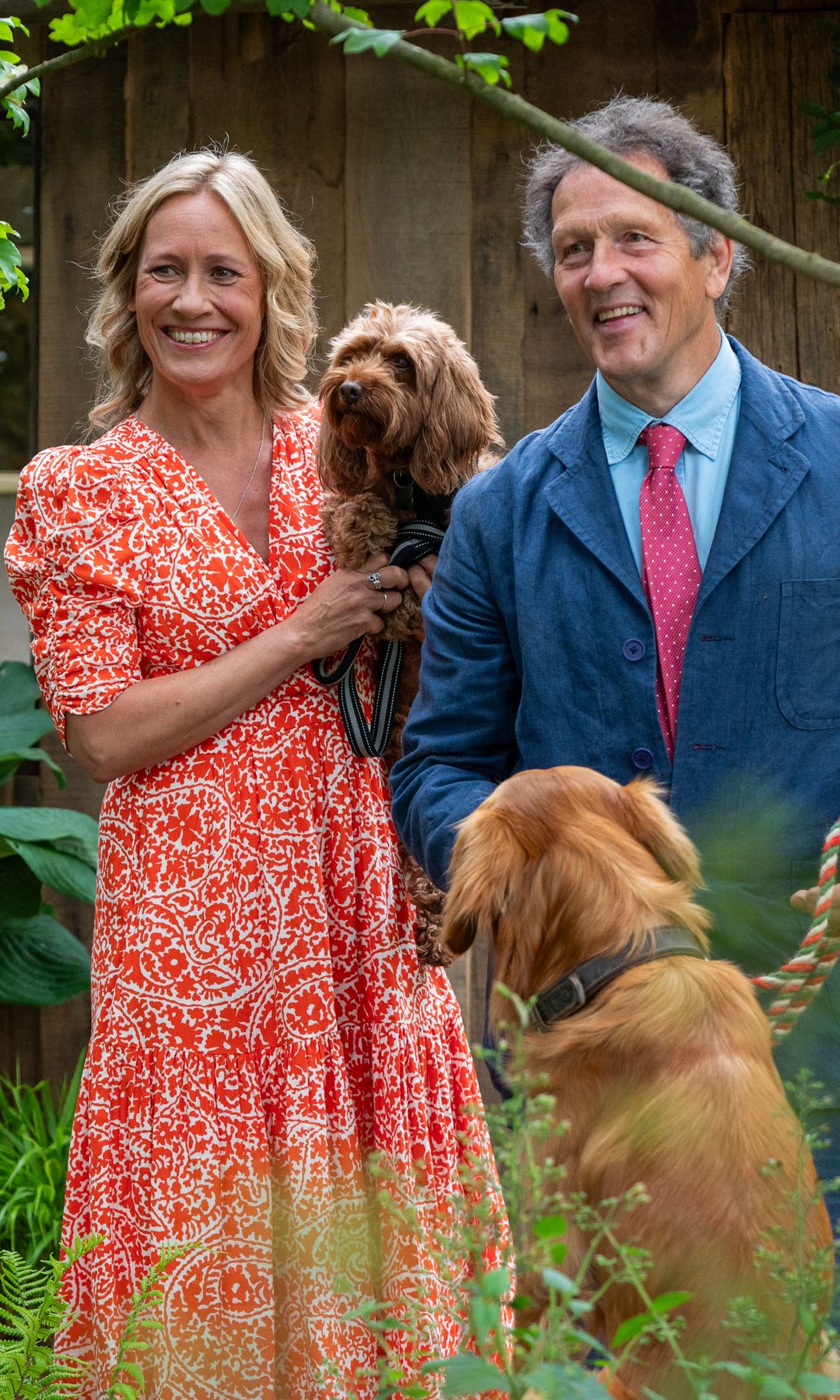Sophie Raworth in a red patterned dress and Monty Don in a blue blazer pose in a garden with two dogs in tow.