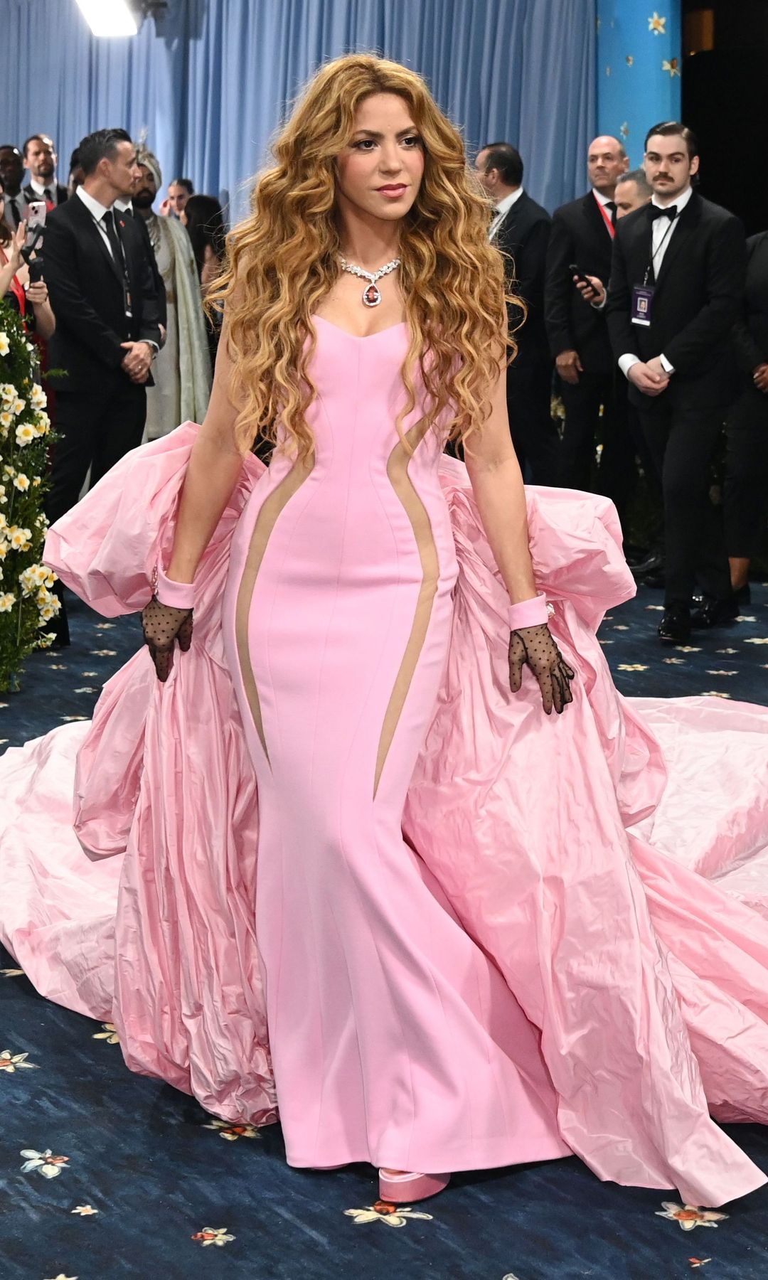 Shakira wears a bubblegum pink gown with huge train detail at the back. She wears her hair in loose beachy waves and is wearing short black mesh gloves.