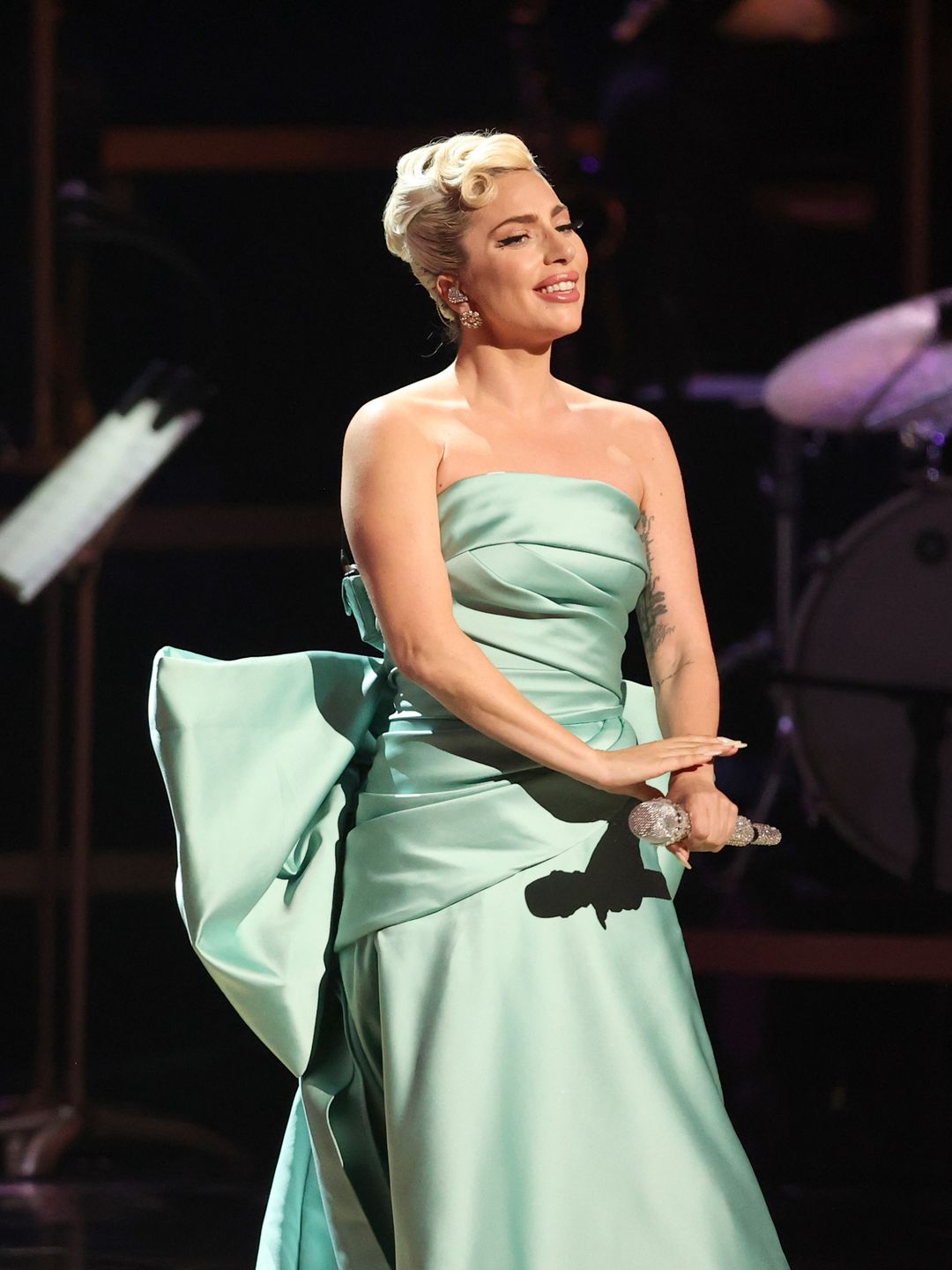 Lady Gaga stands on stage at the 2022 Grammy awards with a chic chignon up do and flawless makeup. She is wearing a mint green strapless dress with a big bow on the back and is holding a microphone.