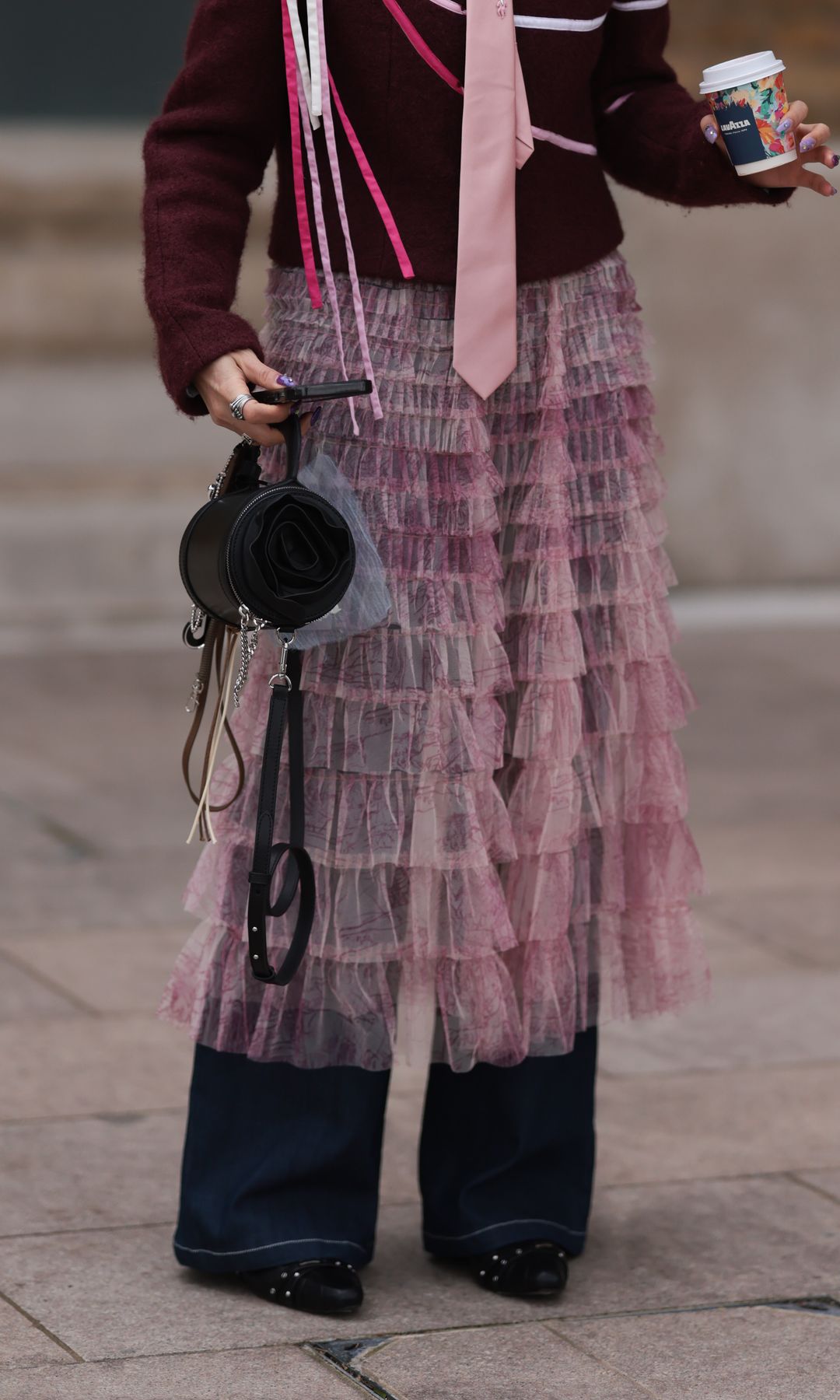  Fashion Week Guest seen wearing a striking mix of textures and colors. She sports a burgundy and pink jacket adorned with swirling patterns and white piping details. A pale pink tie with a decorative brooch adds a unique, almost formal contrast to the playful elements of the look. Over their navy-blue trousers, she wears a sheer, ruffled pink skirt, giving an avant-garde layering effect. Accessories include a small structured black bag with a rolled fabric detail, futuristic cat-eye glasses, and a small, embellished hat with a geometric patch on the front. The ensemble combines eclectic and structured elements, creating a bold fashion statement outside Emilia Wickstead Show during London Fashion Week February 2025