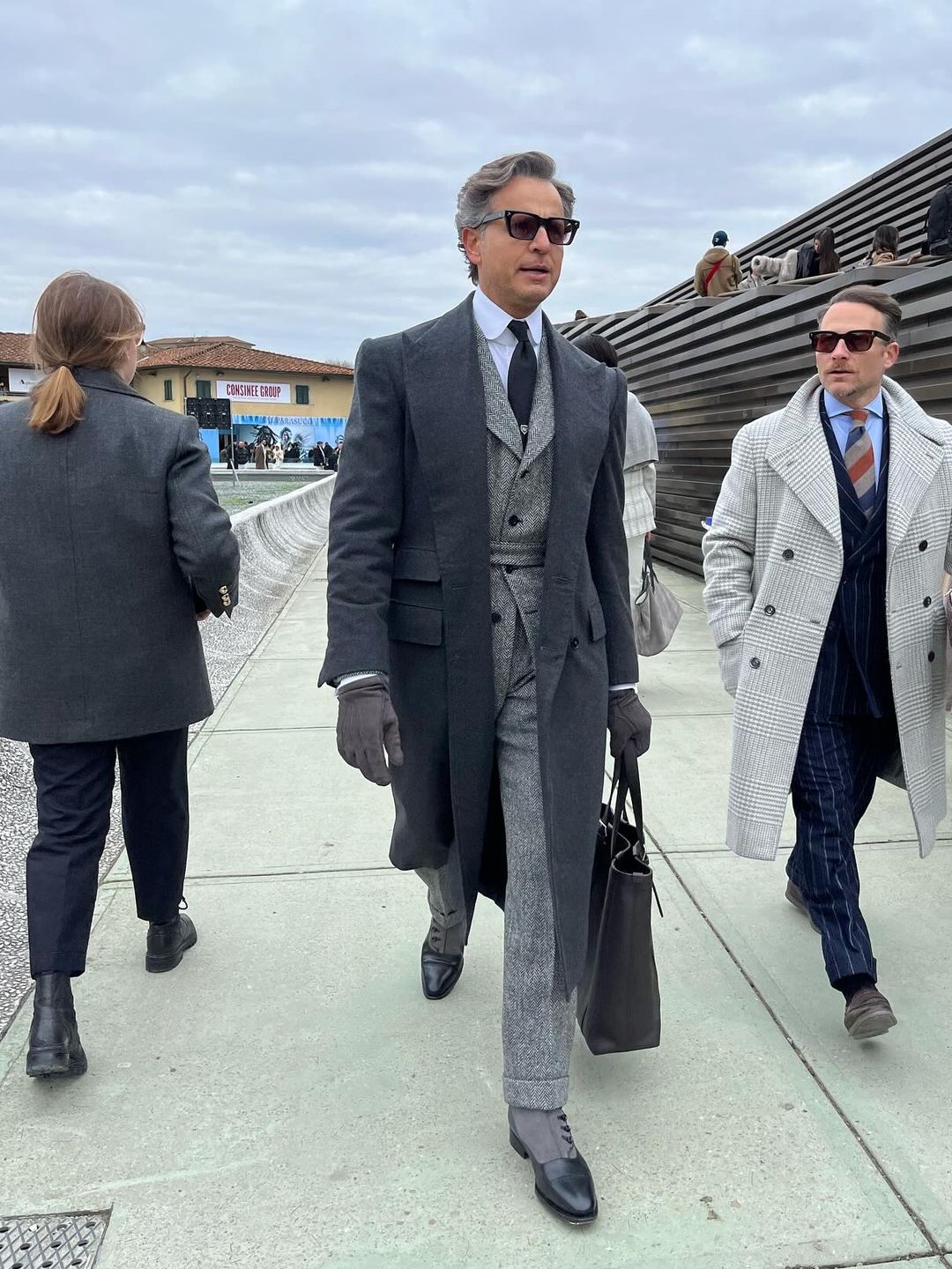 Wearing a grey suit, overcoat, gloves and leather tote bag, this seriously chic guest attends Piti Uomo