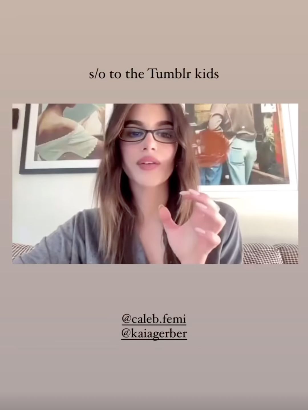 The image features Kaia Gerber, a model and actress, sitting in a well-lit, stylishly curated space. She is wearing small, rectangular-framed glasses with a subtle tint, which rest elegantly on her face. Her long, chestnut brown hair falls in soft waves around her shoulders, and she is dressed in a relaxed, gray knit top. Behind her, a large framed artwork features a scene of people in casual clothing, adding a modern yet artistic backdrop to the setting. Gerber is captured mid-sentence or mid-gesture, her hand raised slightly as if emphasizing a point. The overall aesthetic of the image is sophisticated yet effortlessly cool, highlighting her signature understated elegance.