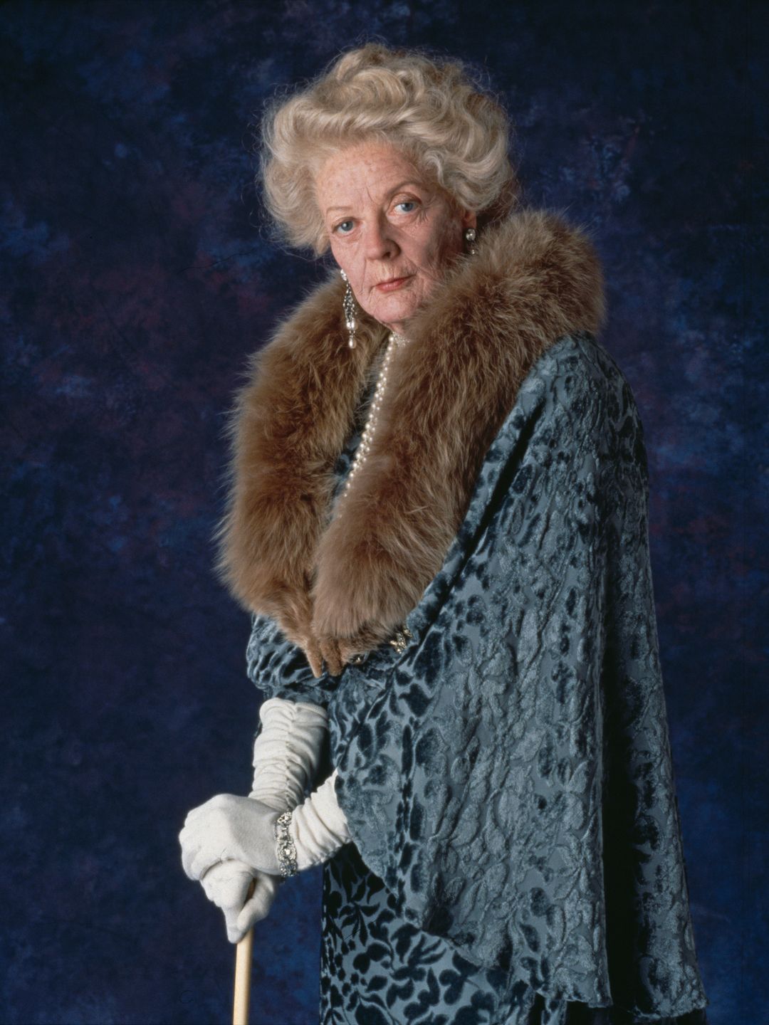 British actress Maggie Smith on the set of Spielberg's film Hook, Maggie Smith stands dressed in an opulent, old-fashioned outfit with a fur collar draped around her neck. 