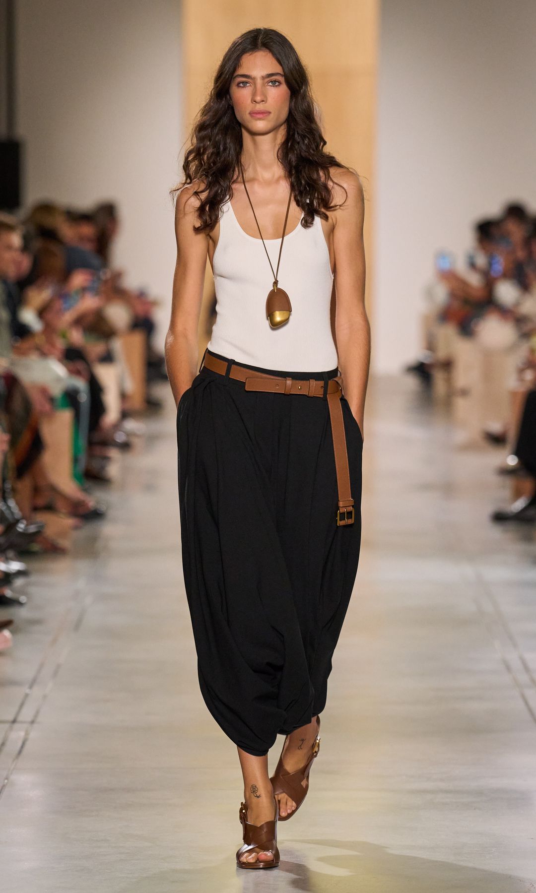 A model at Michael Kors wearing a white vest, long pendant necklace, black oversized trousers and brown belt and sandals