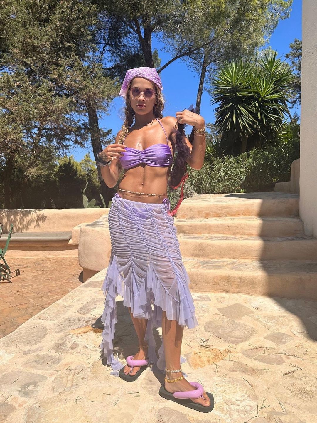 Rita Ora wearing a lilac holiday outfit including a bikini top, sheer skirt, bandana and padded flip flops