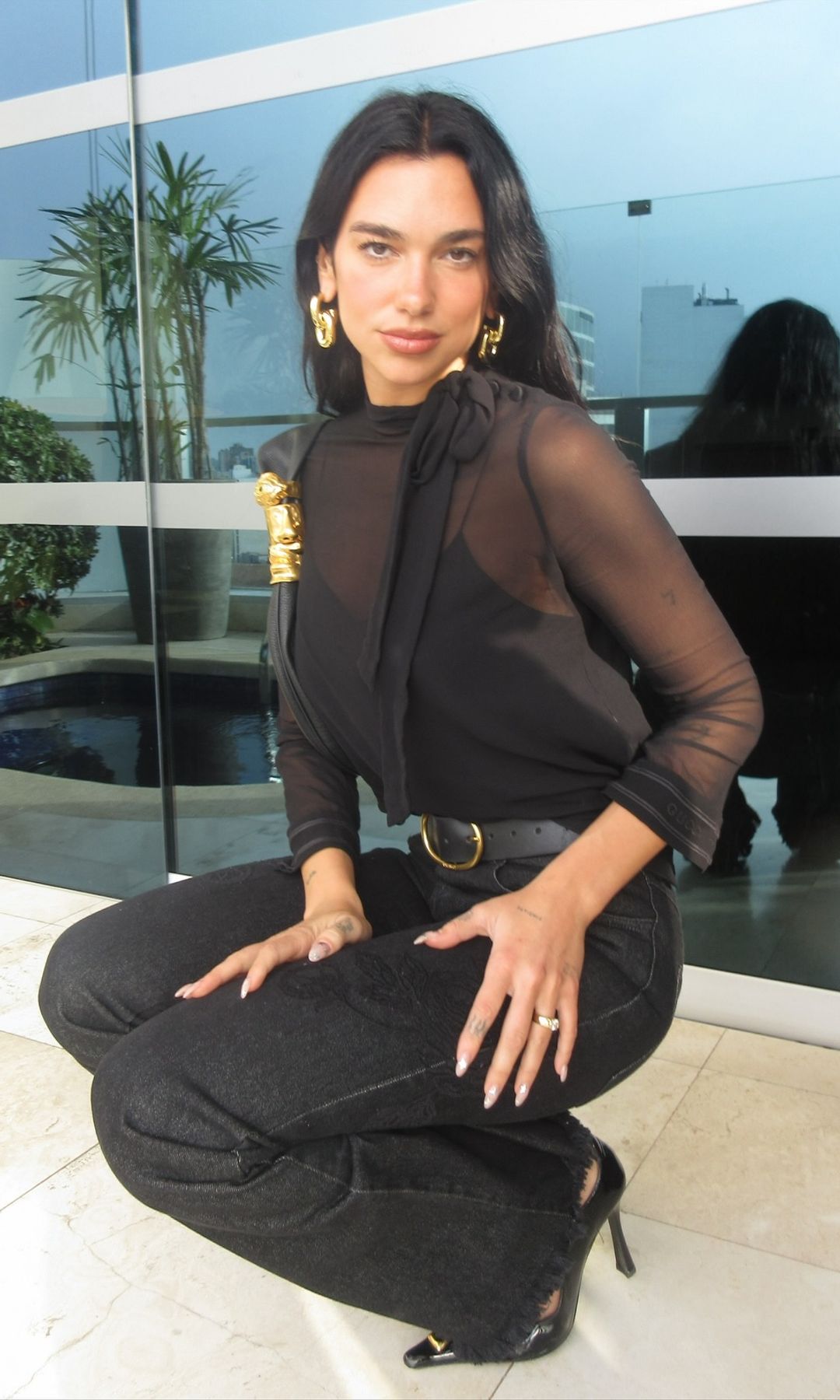 Dua Lipa poses for a photo crouched down in black jeans and a sheer black top