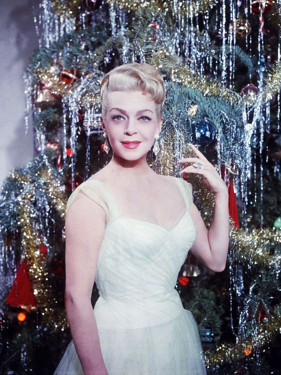 This is a color photograph featuring a woman posing in front of a beautifully decorated Christmas tree. She has light hair styled in an elegant, vintage updo and is wearing a glamorous, light-colored evening gown with sheer straps. The tree behind her is adorned with shiny silver tinsel, gold garlands, and various colorful ornaments, creating a festive and sparkling backdrop. The woman smiles confidently, with one hand raised near her face, showing off a ring and earrings that add to her sophisticated appearance. The scene exudes a classic, holiday celebration vibe.