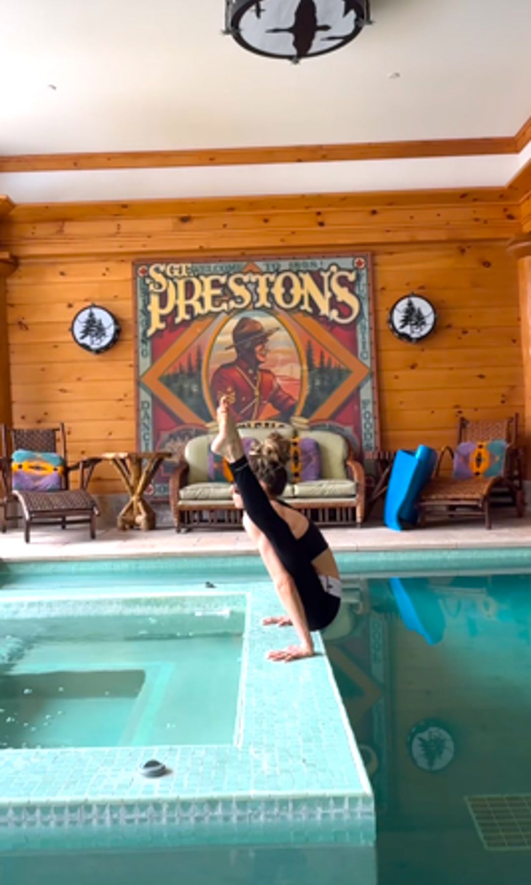 Stacey Bendet balances in a yoga pose at the edge of an indoor pool, crouched like a poised ballerina. She wears black athletic wear, hair piled in a high bun. The rustic wood interior and vintage “Preston’s” sign set a cozy lodge vibe.