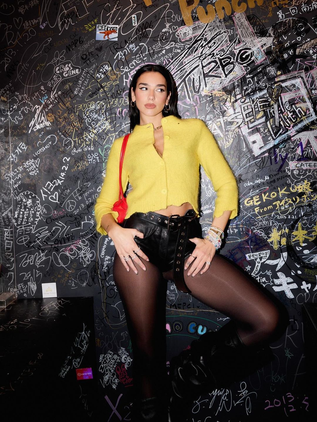 Dua Lipa poses in a yellow knitted cardigan, leather hot-pants and sheer tights