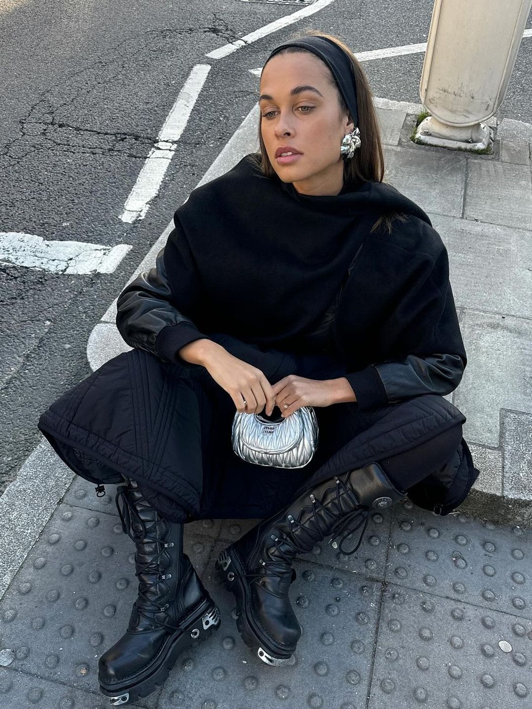 Sarah Lysander wears an all black outfit to sit on a curb in London and accessorises her outfit with a silver metallic Miu Miu bag and bold earrings 