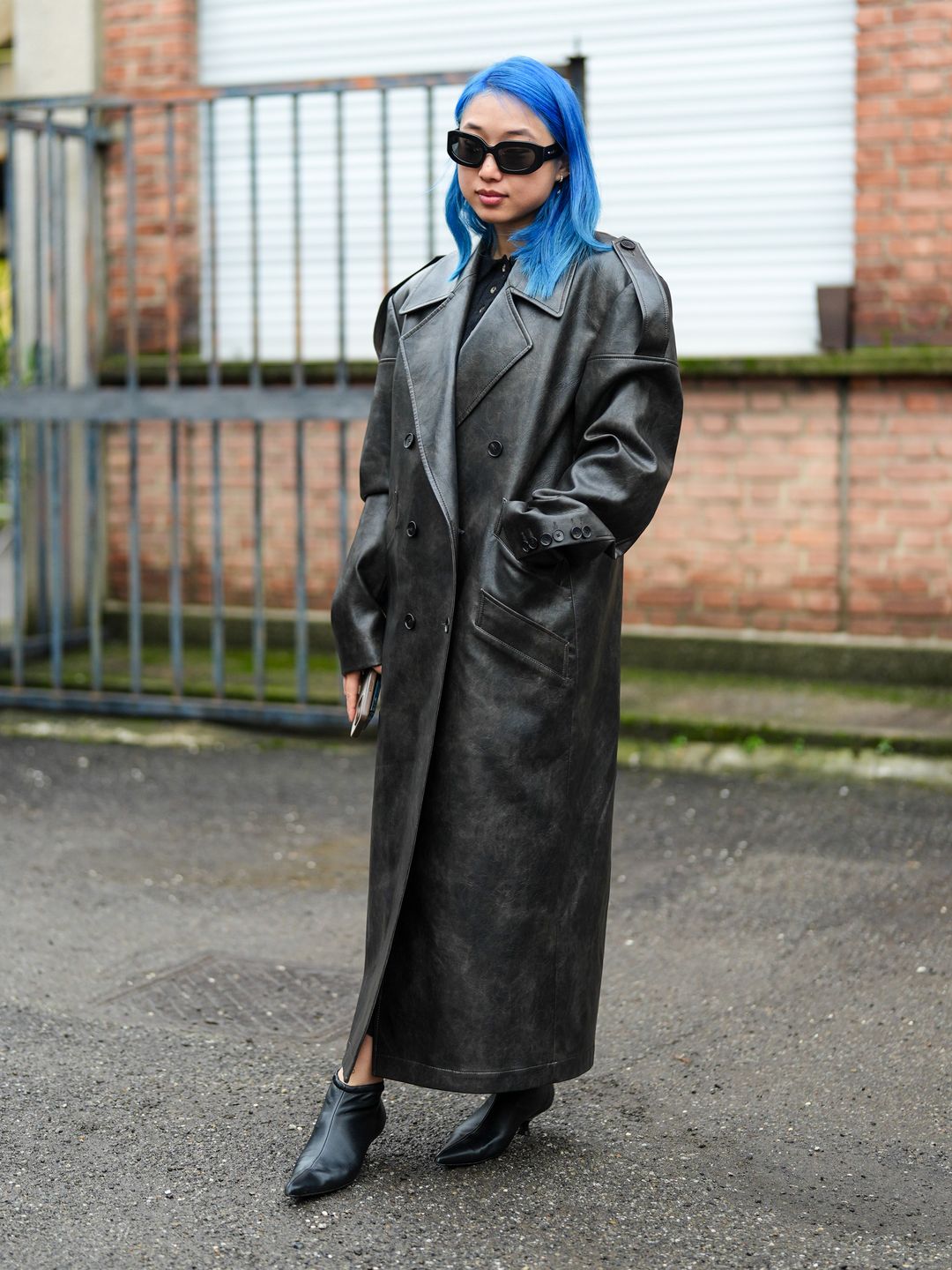 Margaret Zhang wears sunglasses, a black leather double breasted trench long coat, kitten heels black leather pointed shoes outside Gucci's F/W 24 show