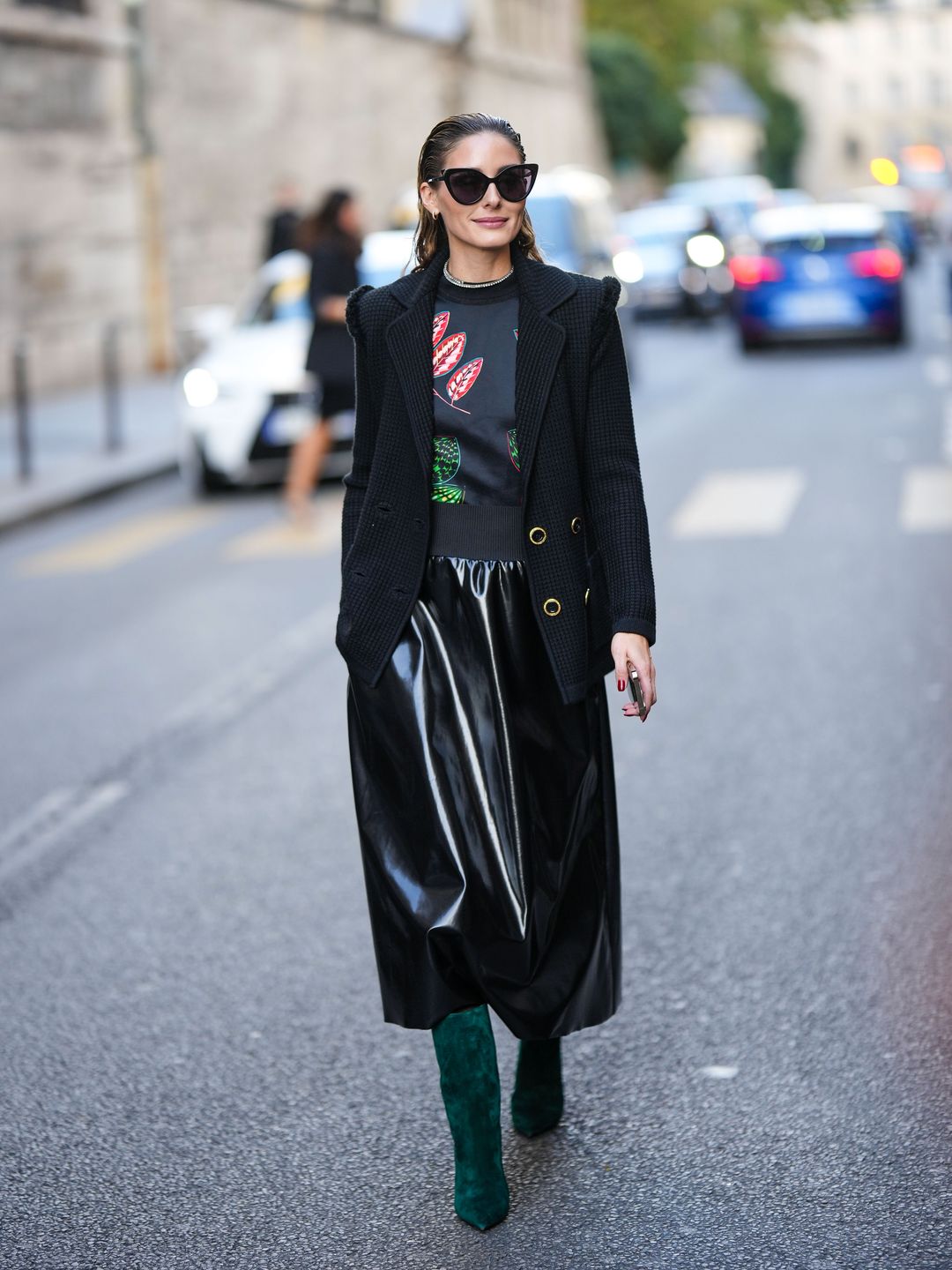 Olivia Palermo on a city street wearing oversized black sunglasses, a black coat with gold buttons, a graphic print shirt, a shiny black midi skirt, and green boots.