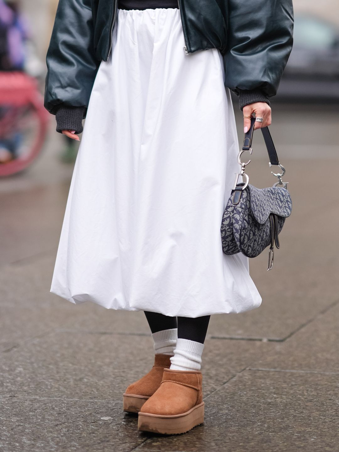 A guest wears sunglasses, a colored scarf, a green leather bomber jacket, a black top, a midi white skirt, white socks, brown suede Ugg shoes, outside Aeron,  during the Copenhagen Fashion Week AW24