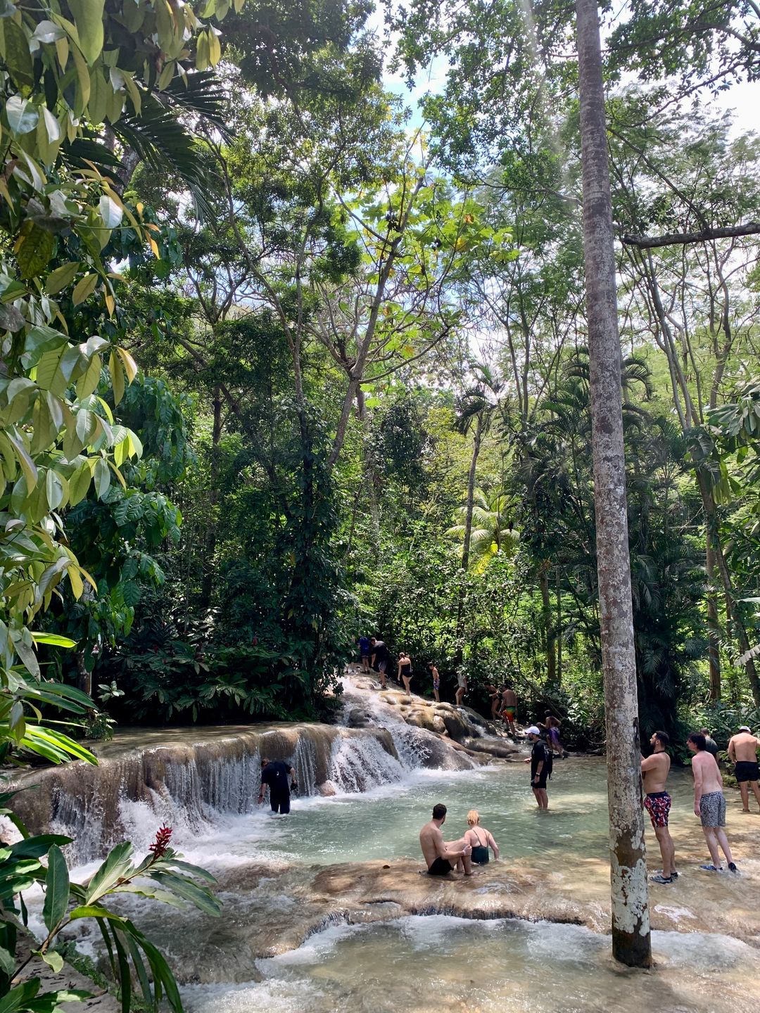 A scenic tropical waterfall surrounded by lush green foliage, with people wading, climbing, and swimming in the cascading pools.