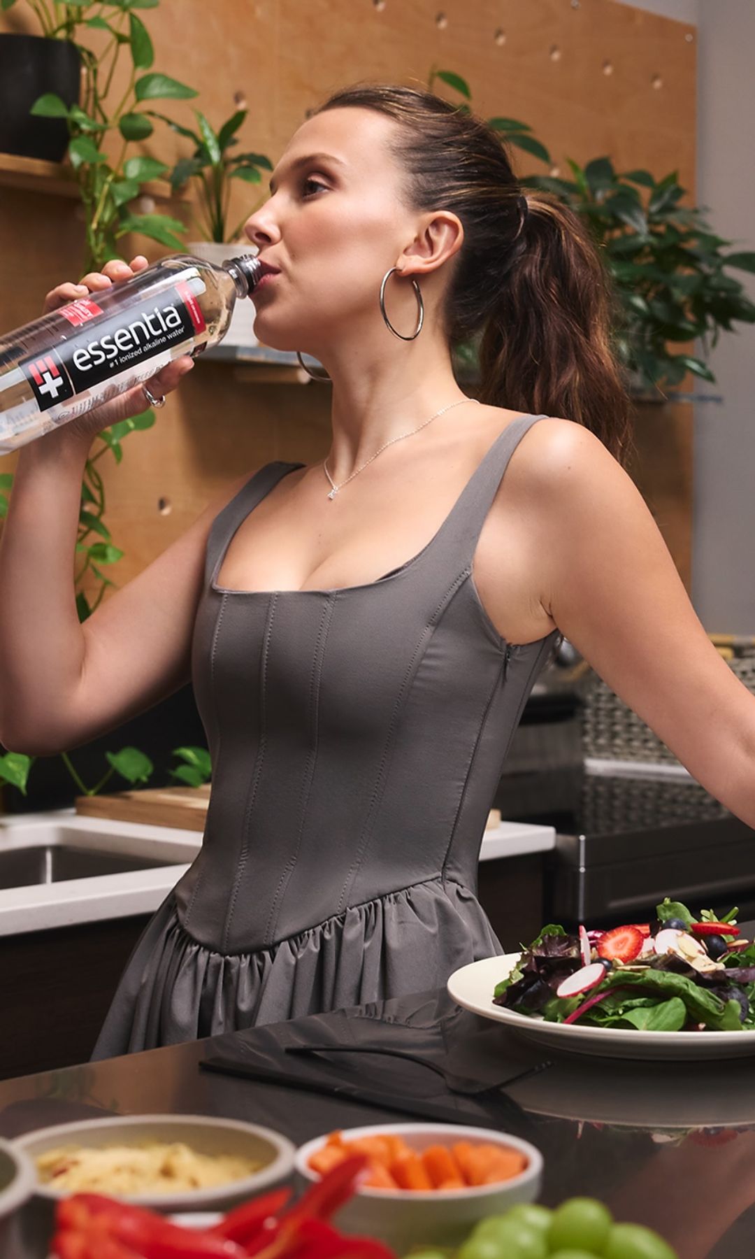 Millie Bobby Brown is photographed wearing a grey mini dress drinking a bottle of water in her kitchen 