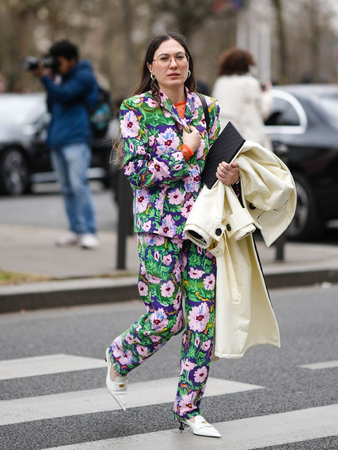 A guest wears silver earrings, a purple with pale pink / green / yellow flower print pattern blazer jacket, matching purple with pale pink / green / yellow flower print pattern suit pants, a pale yellow long coat, white shiny leather pointed heels loafers from Miu Miu , outside Miu Miu, during Paris Fashion Week