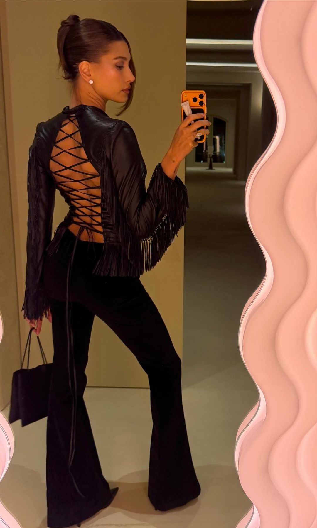 Hailey Bieber poses in a mirror wearing black trousers and a leather jacket